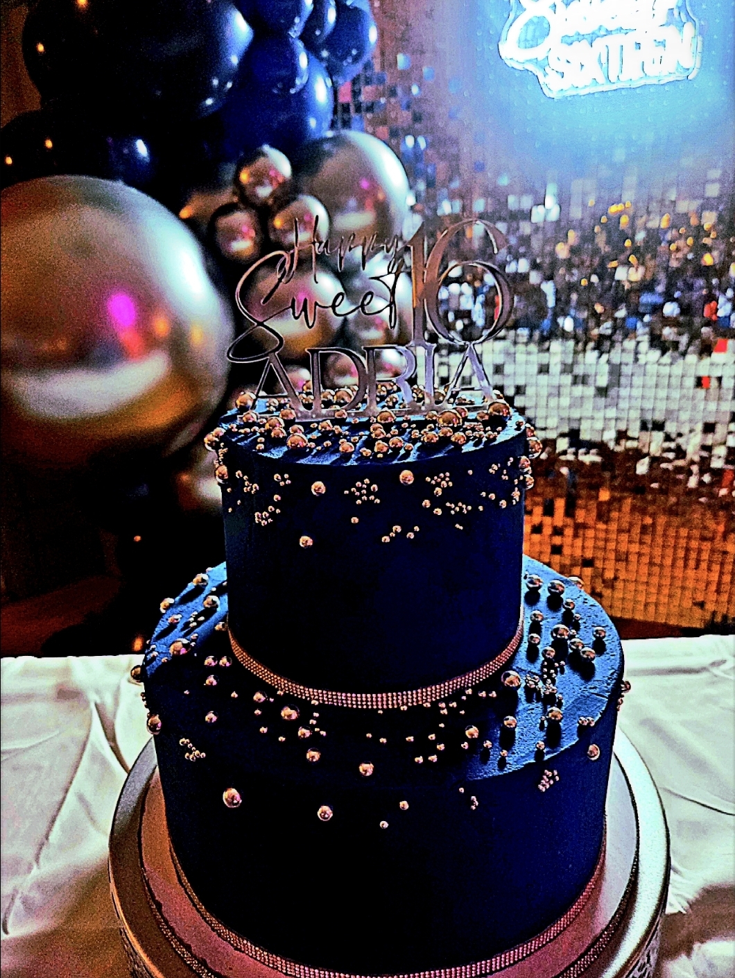 Navy and rose gold two-tier Sweet 16 cake for Adria with balloon garland and neon Sweet Sixteen sign in background