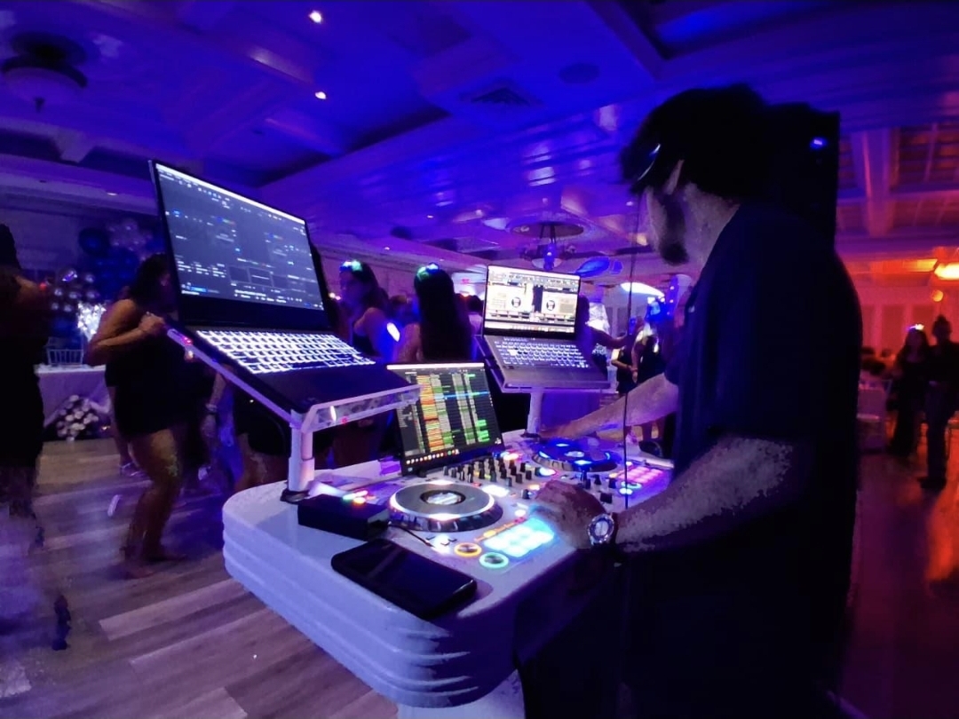DJ Aiden Jules mixing at a full white DJ booth with deep blue uplighting and guests dancing behind him