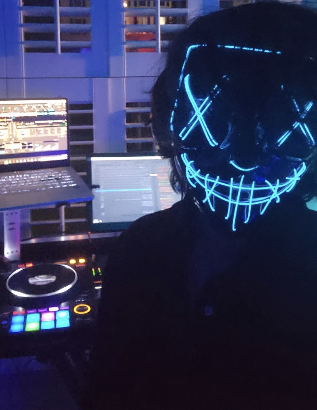 DJ in a glowing blue LED mask performing at a private party with laptop and controller lit by stage lights