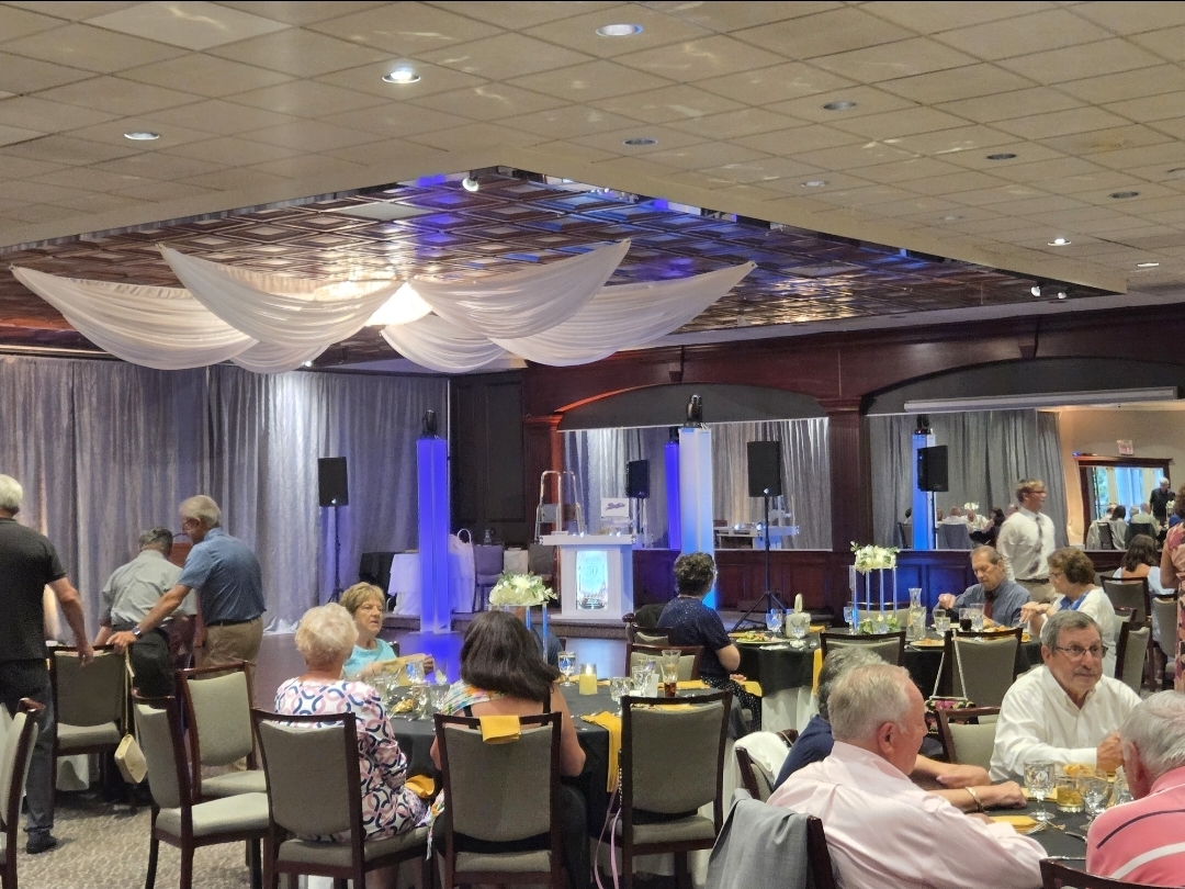 Guests seated at round tables during a 50th anniversary celebration with draped ceiling, DJ booth, and blue and purple uplighting