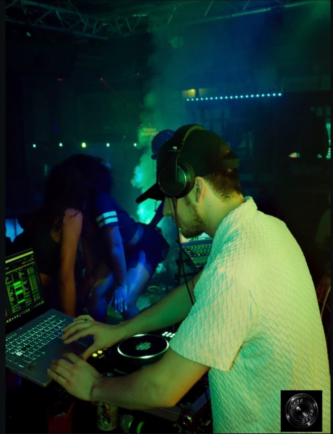 DJ Aiden Jules mixing in a nightclub booth under green haze with a crowd close to the decks