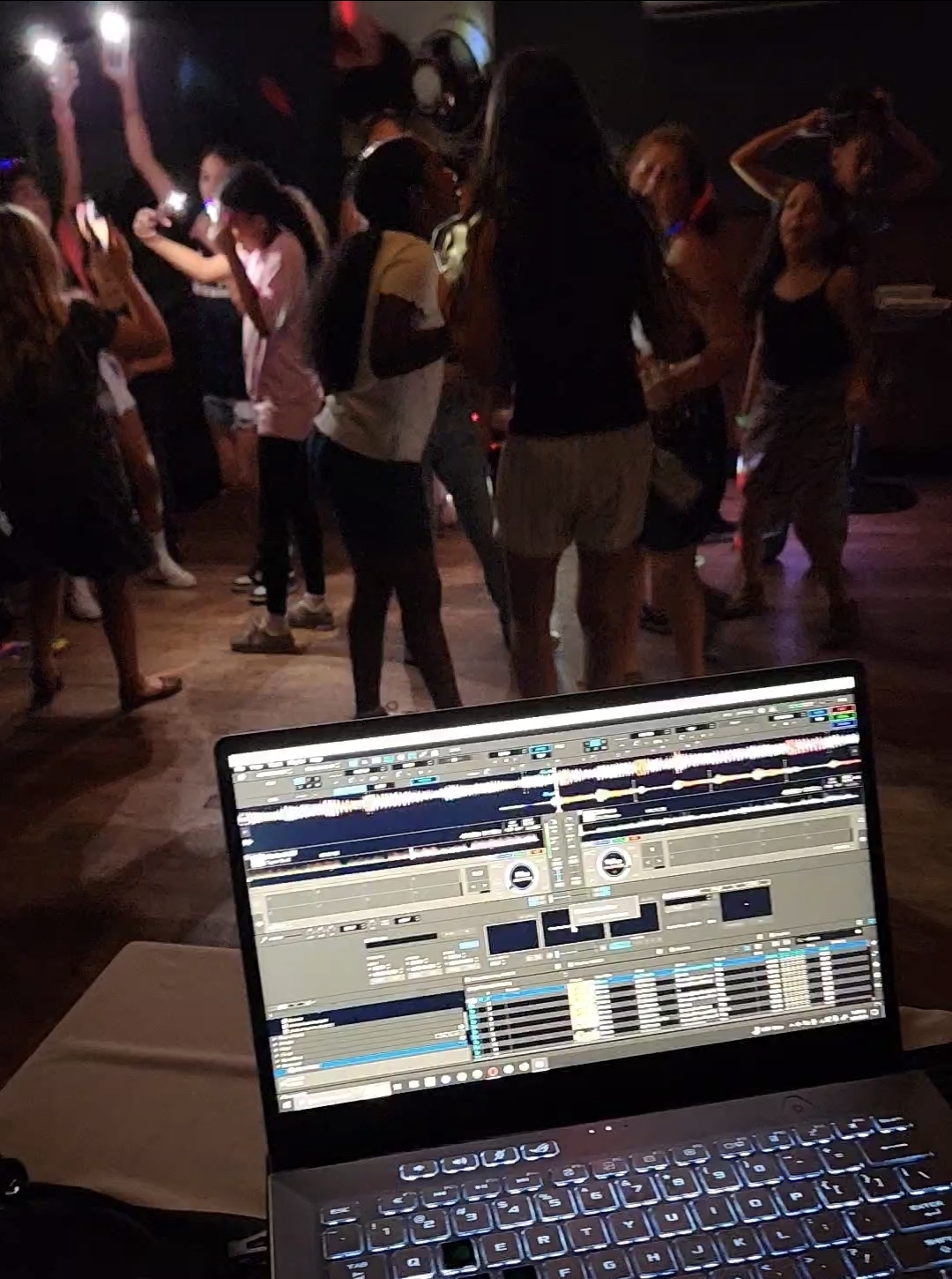 Guests with phones raised on a dark dance floor at a birthday party behind the DJ's illuminated laptop