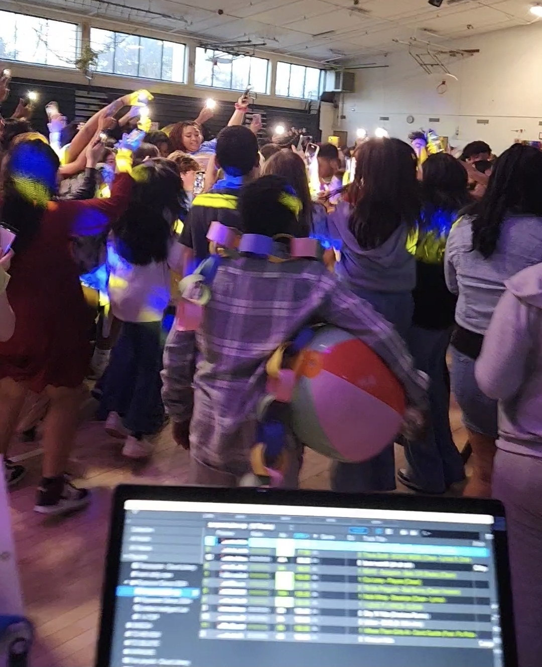 Students dancing under blue and yellow lighting at a school dance with DJ laptop in the foreground
