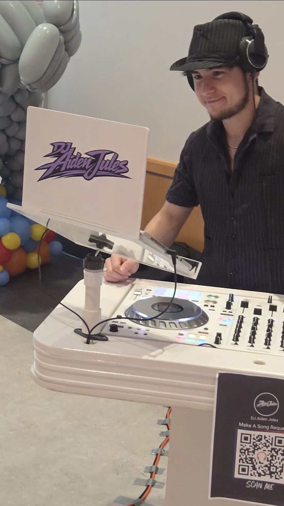 DJ Aiden Jules smiling behind a branded white booth with balloon decor at a private celebration