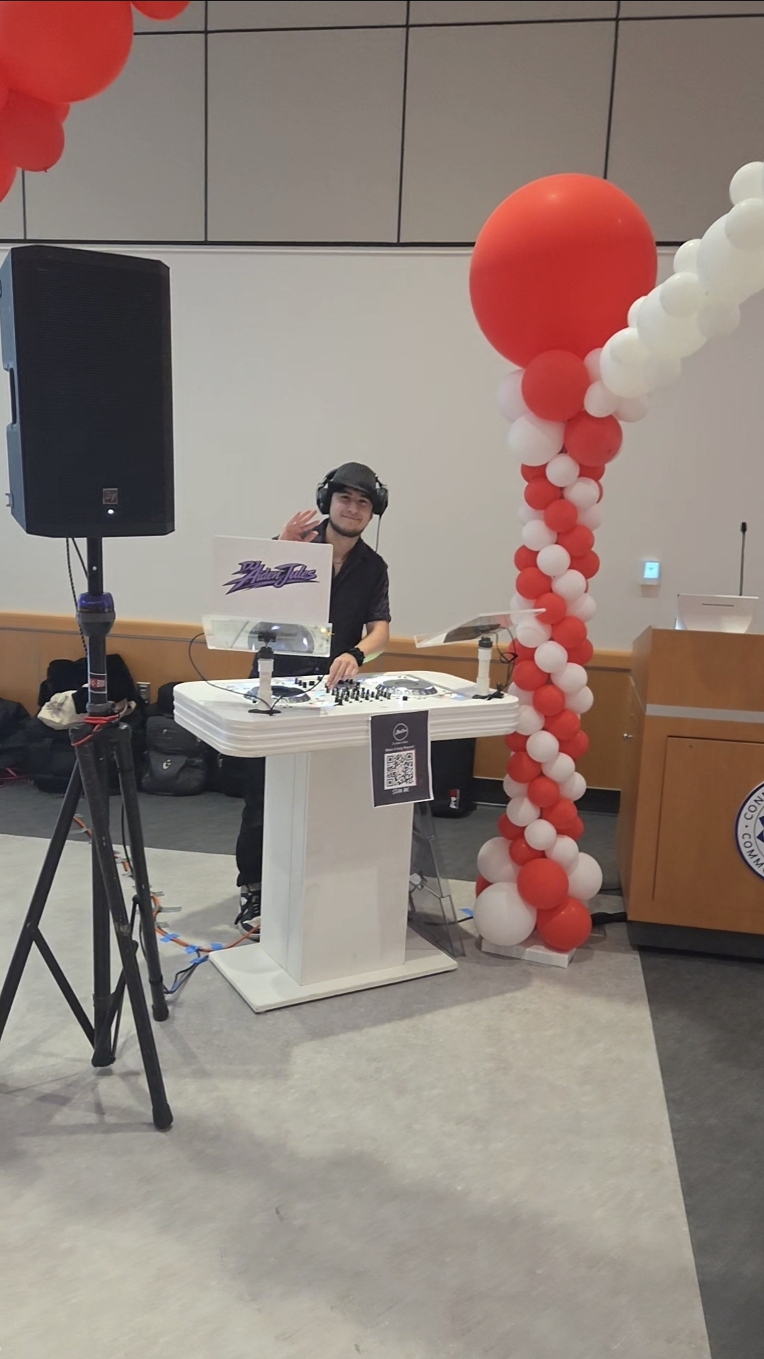 DJ Aiden Jules waving from the DJ booth at a college event with a branded logo panel and balloon column decor