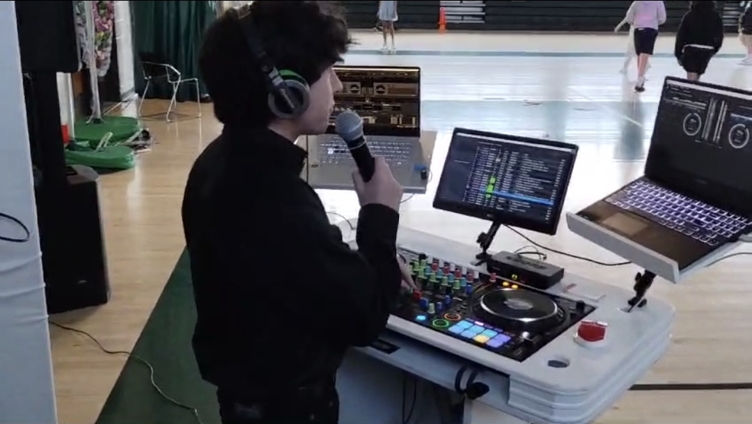 DJ Aiden Jules on the microphone hosting a school-gym birthday celebration with laptop, mixer, and speaker