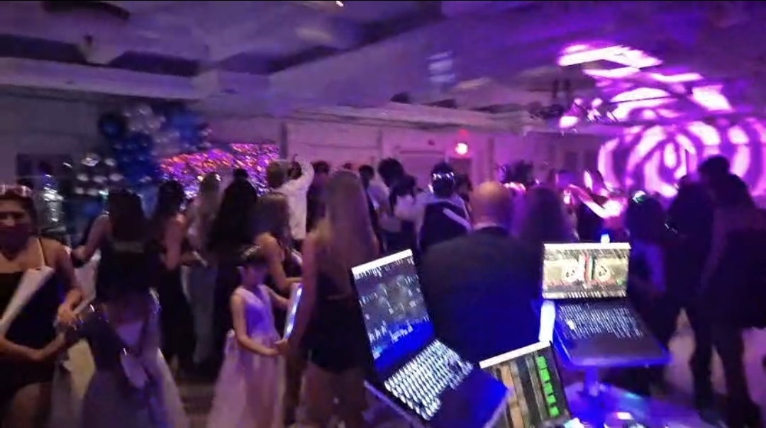 Packed Quinceañera dance floor with purple stage lighting, DJ booth, and guests celebrating