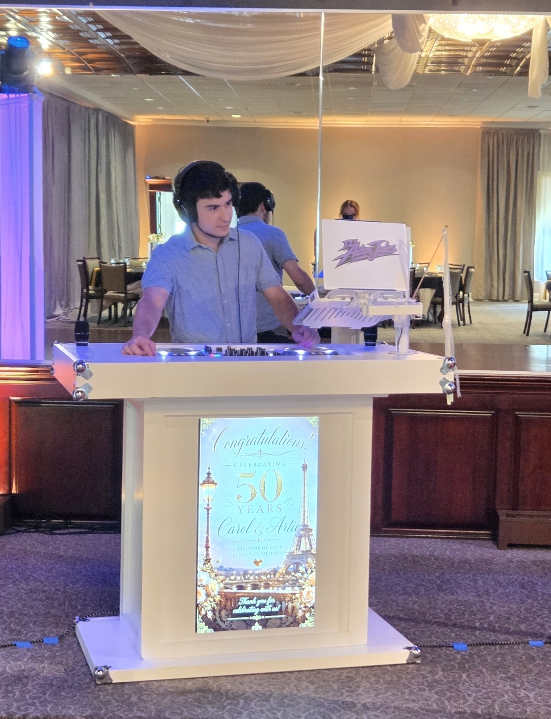 DJ Aiden Jules mixing in headphones behind a white booth featuring a personalized 50th anniversary sign in an elegant ballroom