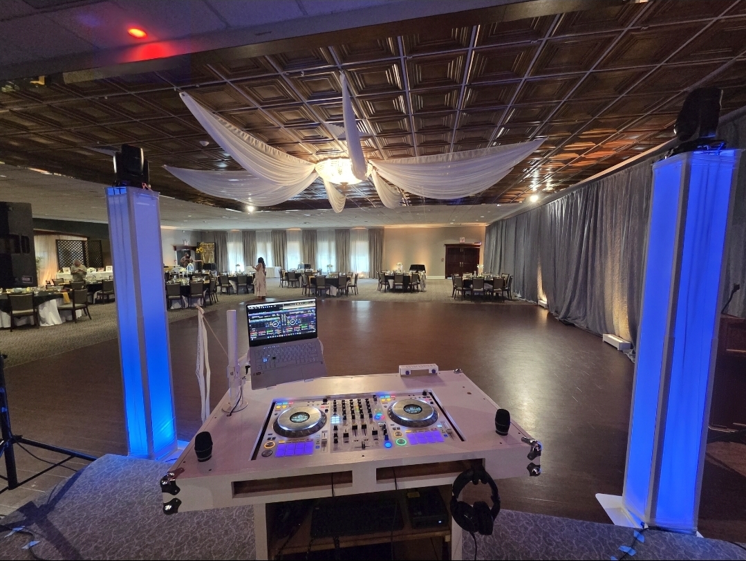 DJ view from behind the booth of an open dance floor with blue LED light towers, draped ceiling, and guests seated at anniversary dinner tables